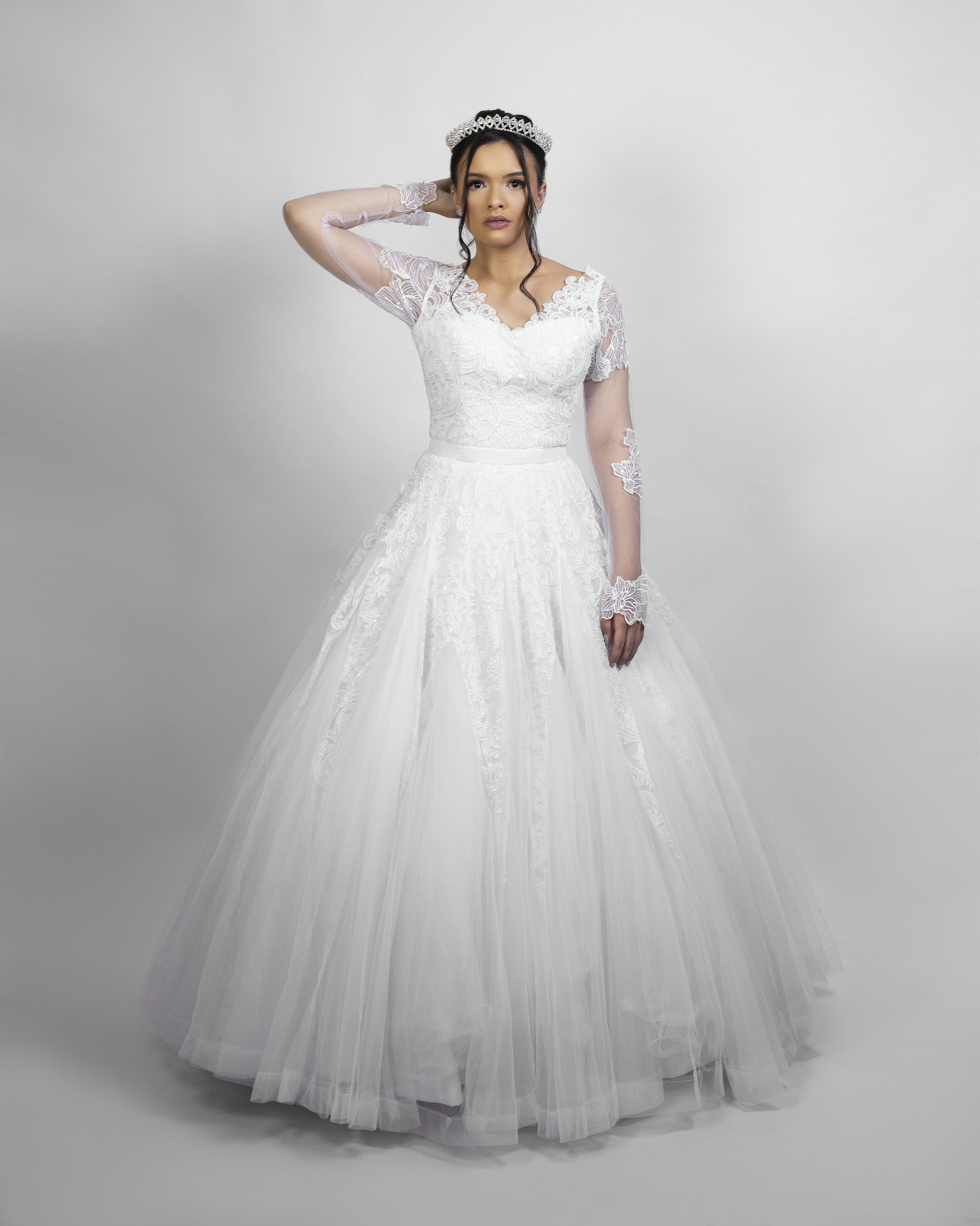 a fulllength studio shot of a bride in a white wedding dress. the dress is a ballgown style with long lace sleeves and a tulle skirt. she is wearing a tiara and gloves. she is standing against a light grey background.