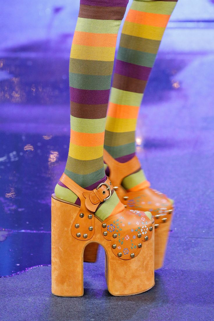 a closeup of a model's legs wearing striped socks and platform shoes on a runway. the shoes are bright orange and have a thick platform and a single strap across the top. the socks are striped in shades of green, yellow, orange, brown, and purple. the background is a vibrant purple.