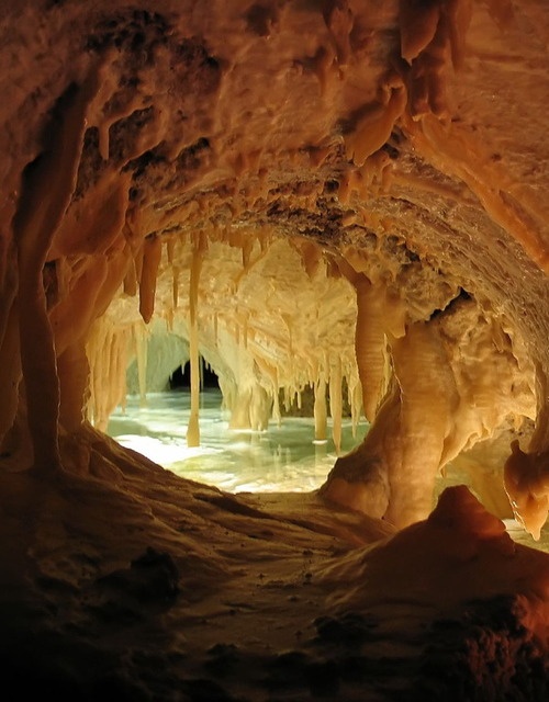 the image shows the interior of a cave with numerous stalactites hanging from the ceiling and stalagmites rising from the ground. there is water present, reflecting the cave's structures and creating a poollike area. the cave is illuminated, highlighting the mineral deposits and textures of the limestone rock formations.