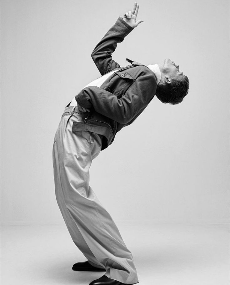 the image is a black and white, fulllength studio shot of a male model striking a dynamic and expressive pose. he is wearing loosefitting, wideleg trousers and a darkcolored jacket. his posture is arched backward, with one arm raised overhead, conveying a sense of movement and energy. the background is plain and uncluttered, drawing all attention to the model's pose and attire. the overall style suggests contemporary fashion photography with an artistic and graceful feel.  the image has a clean, minimalist aesthetic.