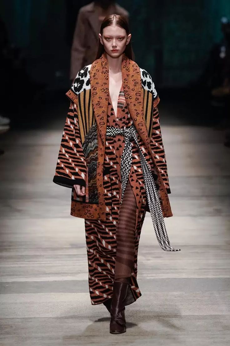 a female model walks the runway in a long patterned dress with black and brown stripes and a large brown and black patterned scarf draped around her neck. the dress has a slight vneck and the model has a belt tied around her waist. she is also wearing a pair of brown boots. the runway is dark and she is in front of a blurry background. the model is posing for the camera.