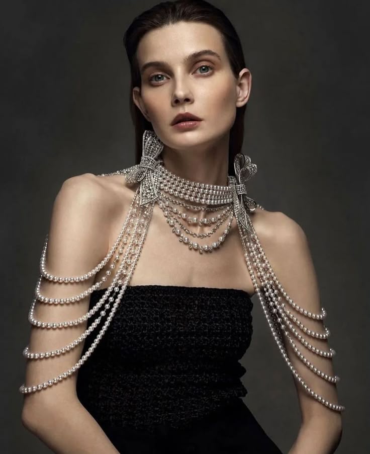 a portrait of a woman wearing an elaborate pearl and diamond jewelry set with a black strapless dress. the jewelry includes a choker with diamond bows on the sides and cascading strands of pearls that drape over her shoulders. the woman has a neutral expression and is positioned against a dark gray background.