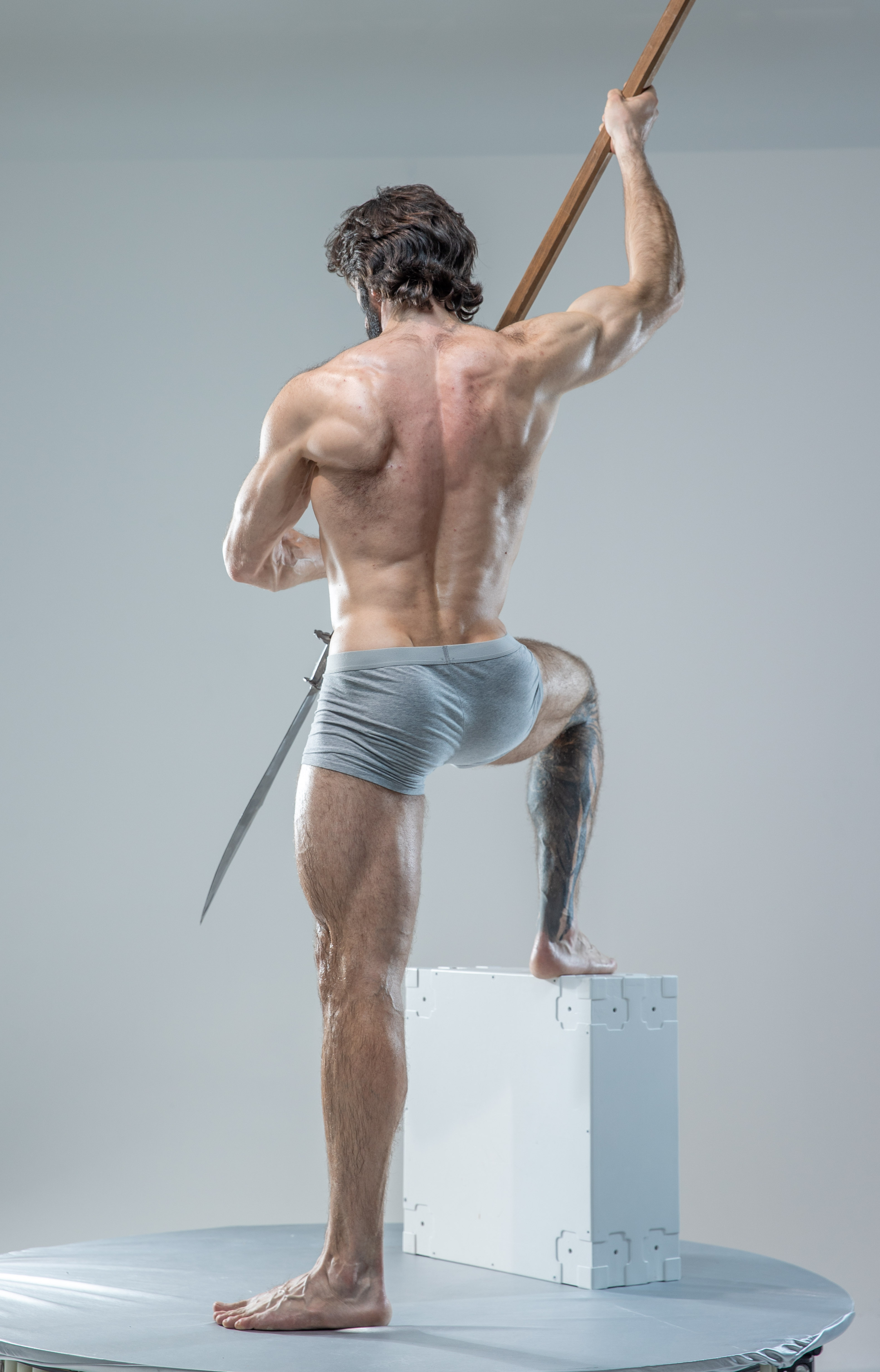 a muscular man with long hair and beard stands in a dynamic pose, his back to the camera. he holds a sword aloft in his right hand, the blade curving down towards the floor. he is shirtless, wearing only grey underwear. he stands with one foot on a white block, which is placed on a round platform. the background is a neutral grey. the image appears to be an art reference photo, showcasing the man's musculature and the dynamic pose.