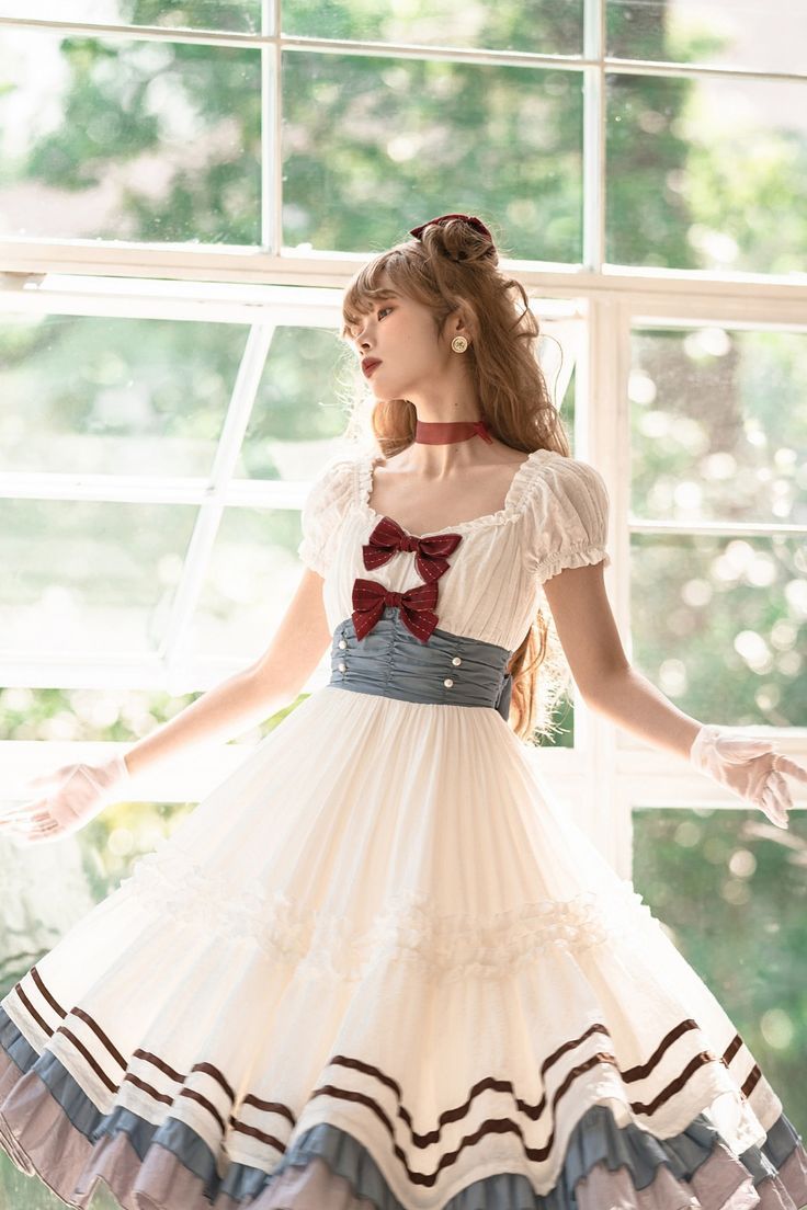 a young woman wearing an elegant white lolita fashion dress stands posing in front of large windows. she has her arms outstretched and head tilted back. the dress has a tiered skirt with blue and brown accents, and a burgundy bow at the waist. the bright, natural light from the windows illuminates the scene.