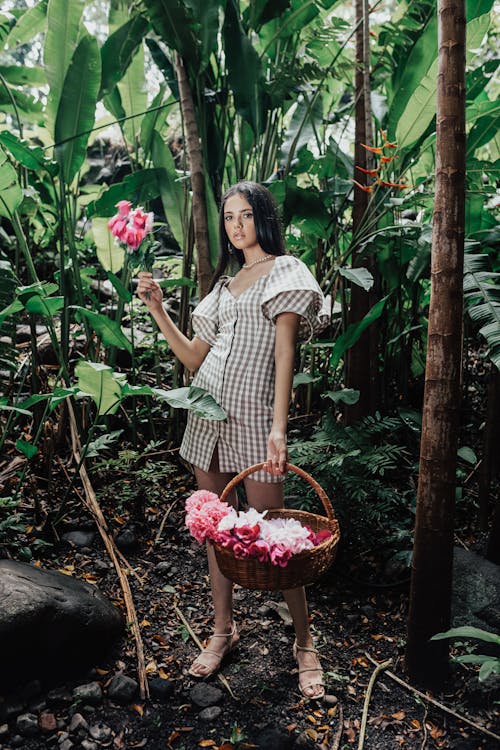 a fulllength shot features a woman standing in a lush, tropical forest, holding a pink flower in her left hand and a woven basket filled with pink and white flowers in her right. she is wearing a short, checkered dress with puffy sleeves and tan sandals, and is adorned with a necklace. the surrounding forest is dense with large green leaves and tall trees, creating a vibrant and verdant backdrop.