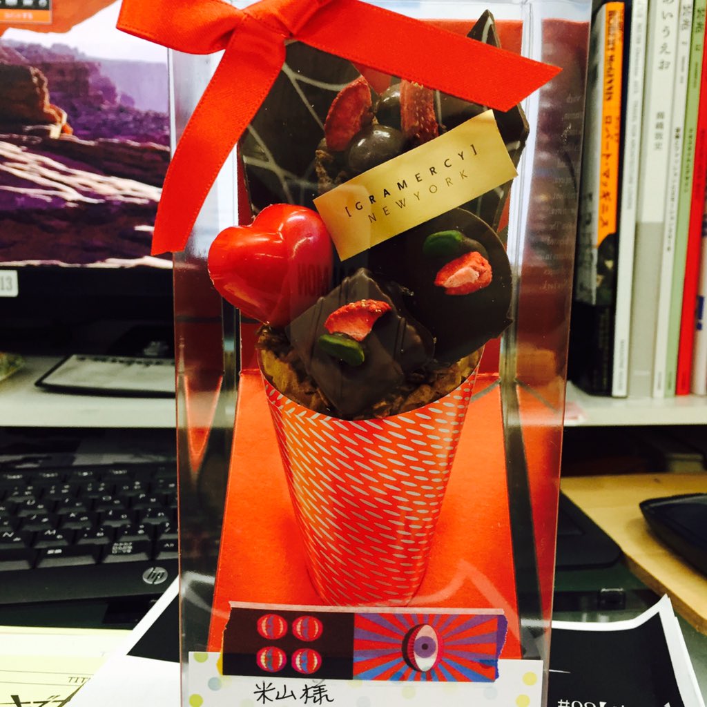 a clear plastic box tied with a red ribbon contains a cone filled with assorted chocolates and a chocolate heart. the cone is decorated with a red and white pattern and sits on a red card. the box is on a desk with a keyboard, books, and a computer monitor in the background.