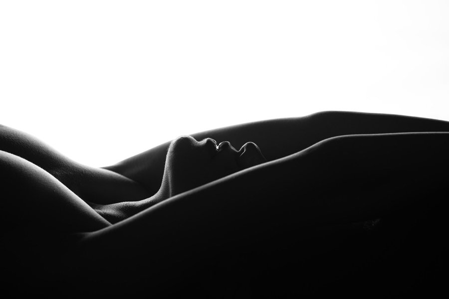 the image is a stark, black and white silhouette of a reclining female nude.  the body is positioned with arms outstretched above the head, creating smooth, flowing lines against a bright white background.  the focus is on the form and curves of the body, with the absence of detail emphasizing the abstract and sensual nature of the image.  the strong contrast between light and shadow enhances the dramatic effect.  the overall aesthetic is minimalist and artistic.