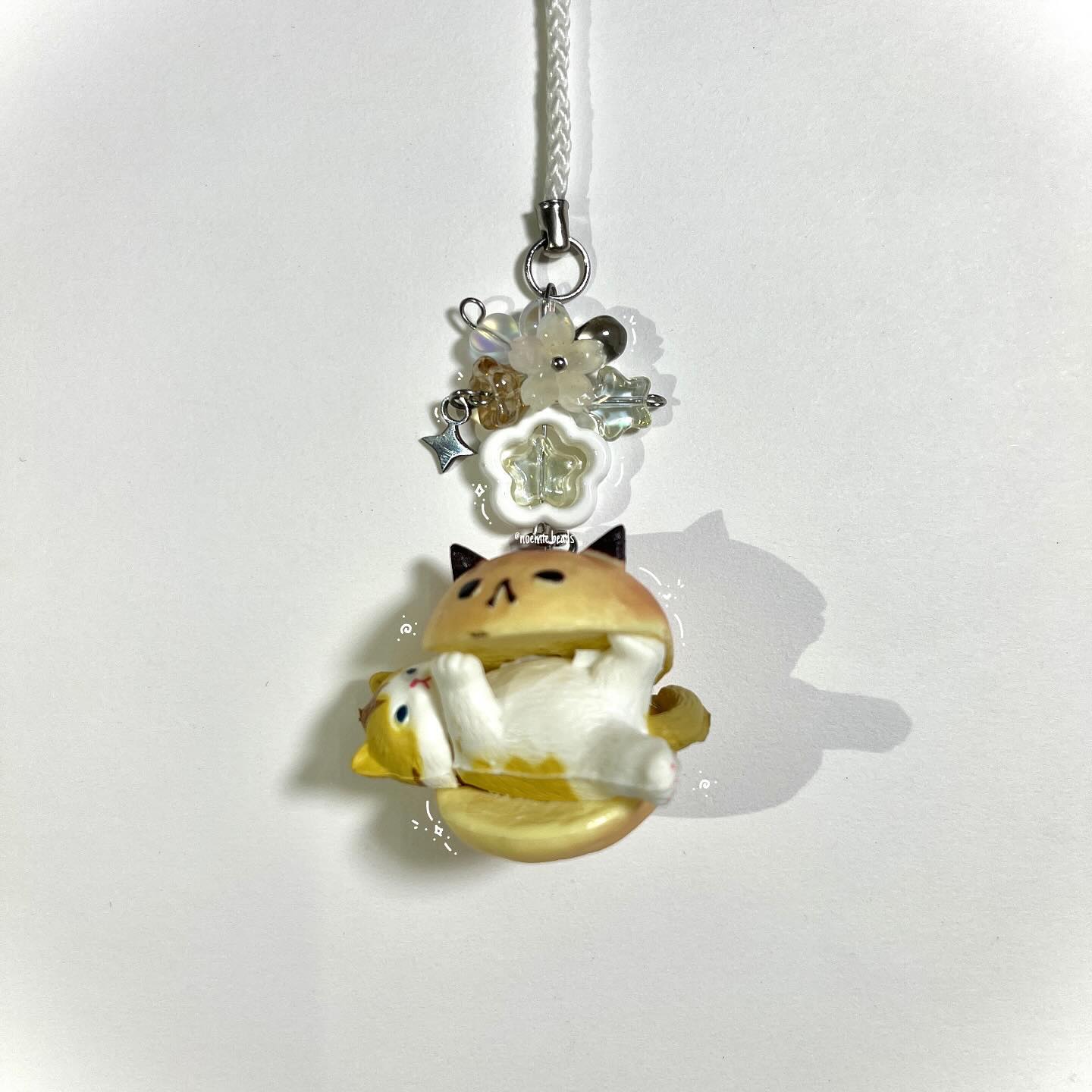a cat themed keychain featuring a cat inside a burger bun and decorative beads and flower attachments hangs from a white string against a white background.