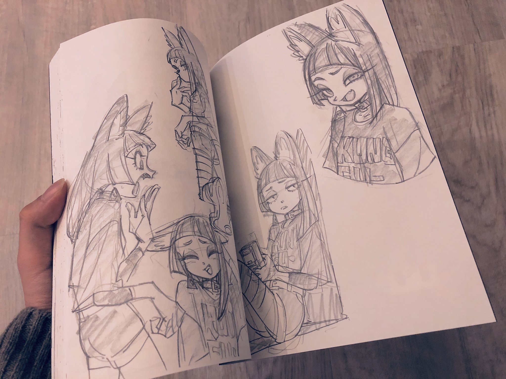 a sketchbook is open to a page with five drawings of an original furry character with fox ears and a tail. she is wearing a band tshirt, choker, and shorts, and has short hair with bangs. the drawings show her in different poses and expressions. the sketchbook is being held open by a hand on a woodlaminate floor. the drawings are done in pencil.