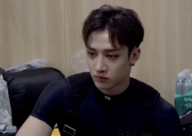 the image shows a closeup of bang chan, a member of the south korean kpop group stray kids. he's seen from the chest up, appearing serious and pensive. he's wearing a black shirt and has a nose piercing and an earring visible. the setting seems to be a backstage or waiting area, indicated by the muted lighting and what looks like luggage or storage containers in the background.  the overall mood is somber and slightly melancholic.