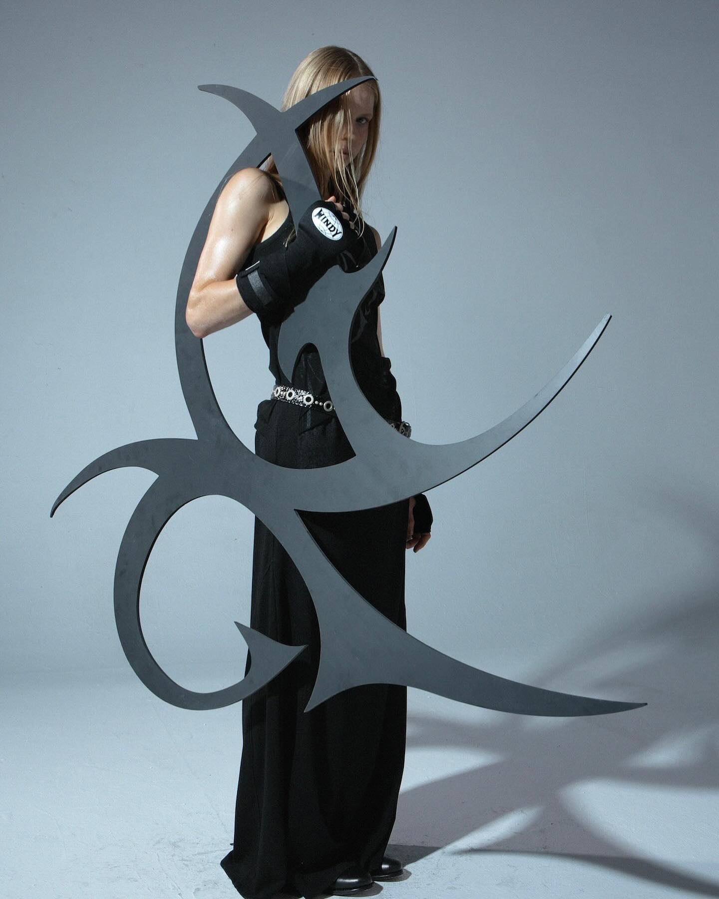 the image shows a female model with long blonde hair wearing a long black dress and black gloves. she is holding a large, abstract, dark gray metal sculpture or weapon that resembles a stylized tribal design. the sculpture is intricately shaped with sharp, angular lines and curves. the model's pose is strong and confident, enhancing the edgy and powerful aesthetic of the image. the background is a simple, light gray studio setting, which draws focus to the model and the dramatic prop. the overall style is dark, moody, and suggestive of fantasy or gothic themes. the gloves are visibly branded as "kindy".