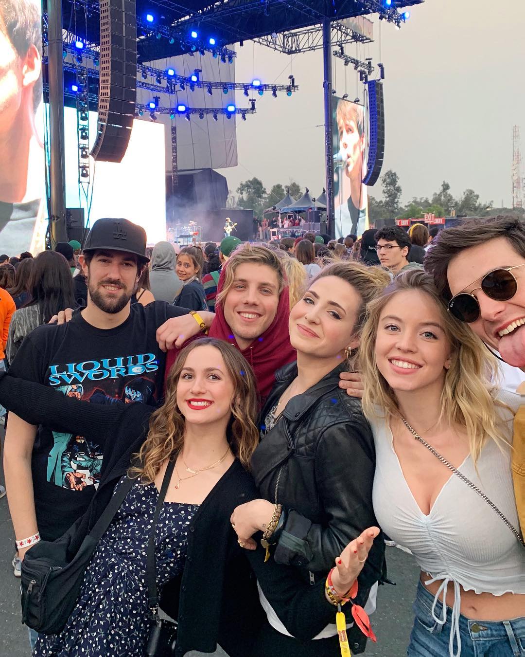 a group of young adults are taking a selfie at a music festival. they are all smiling and looking at the camera. the concert stage is visible in the background. the crowd is blurred, but the lights and energy of the concert are clear. the group consists of both girls and boys and they appear to be friends.