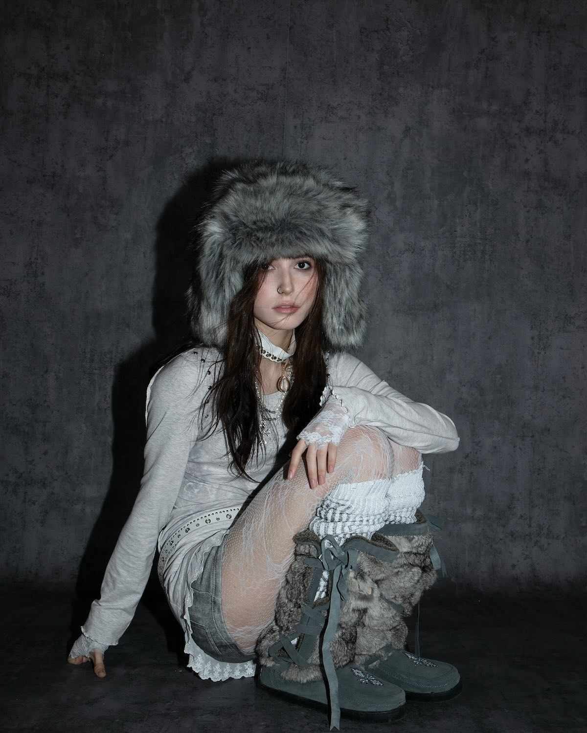 a young woman is sitting on a dark, textured floor against a matching background. she's wearing a gray fur ushanka hat, a light gray longsleeved shirt, gray shorts, and white lacy tights. her footwear is a pair of gray furlined snow boots. her pose is relaxed, and she looks directly at the camera. the overall style suggests a winter or cold weather theme, and the clothing choices are fashionable and somewhat edgy.