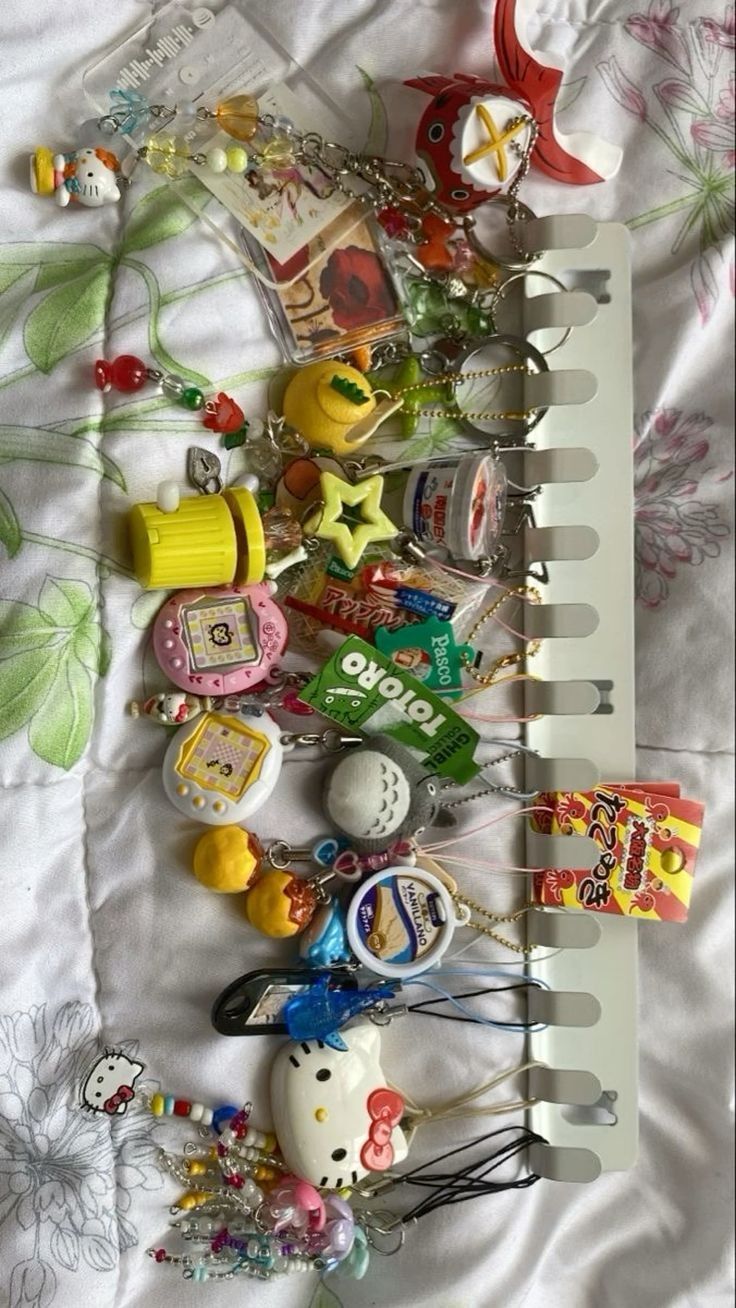 the image shows a collection of keychains and charms hanging on a white display stand, placed on a patterned bedspread. the keychains feature various characters and objects including hello kitty, totoro, tamagotchi, and miniature versions of japanese snacks. there are also other assorted trinkets and accessories.