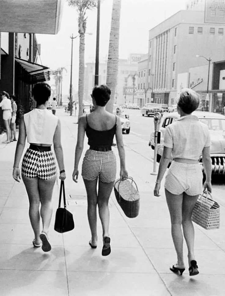 a black and white photograph depicts three women walking away from the camera down a city sidewalk. each woman is dressed in shorts and tops, holding bags or baskets. the woman on the left is wearing patterned shorts and a lightcolored top, while the woman in the middle wears a tank top and gingham shorts. the woman on the right is wearing a shortsleeved shirt and white shorts. the background shows a street scene with cars, buildings, and palm trees.