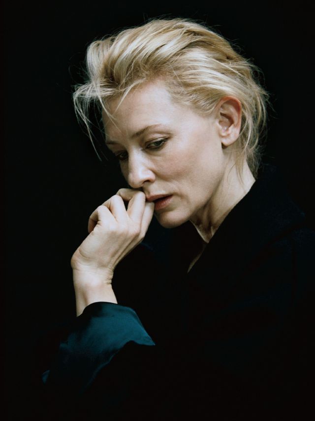 a portrait of cate blanchett with blonde hair against a black background. she has a thoughtful expression, with her hand touching her face near her mouth. the photo is a closeup studio shot, highlighting her serious and introspective mood. she is wearing dark clothing.