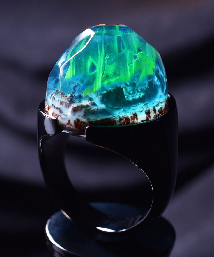 a closeup of a unique ring featuring a large, domeshaped resin cabochon with an abstract design of green and blue swirls, evoking the image of an underwater scene or the aurora borealis. the ring is set in a silver band with a black finish, highlighting the colorful and vibrant nature of the cabochon. the image captures the beauty and intricate detail of the ring, showcasing its potential as a statement piece or a unique gift for someone who appreciates unique and artful jewelry.