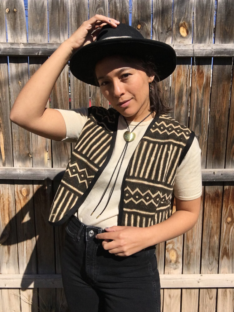 a woman is standing in front of a wooden fence, modeling a black and beige patterned vest over a simple white top and black jeans. she's wearing a black cowboy hat and a long necklace with a heartshaped pendant.  the setting appears to be outdoors, and the sunlight illuminates her. the image showcases a boho or western style fashion, with a focus on the vest's unique design.  the woman's pose and expression are confident and relaxed.