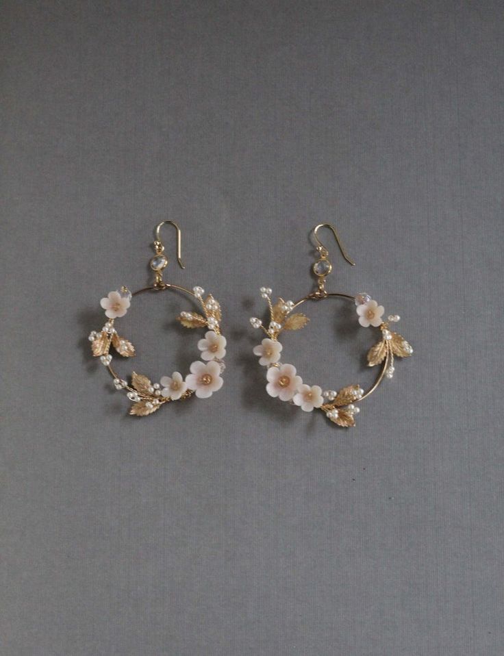 a pair of elegant gold hoop earrings adorned with delicate blush pink flowers and small pearls.  the flowers and leaves are arranged in a wreathlike pattern around the hoop. the earrings are displayed on a gray background, highlighting their detail and delicate nature. they are suitable for wedding or bridal wear, or any occasion requiring elegant and feminine jewelry.
