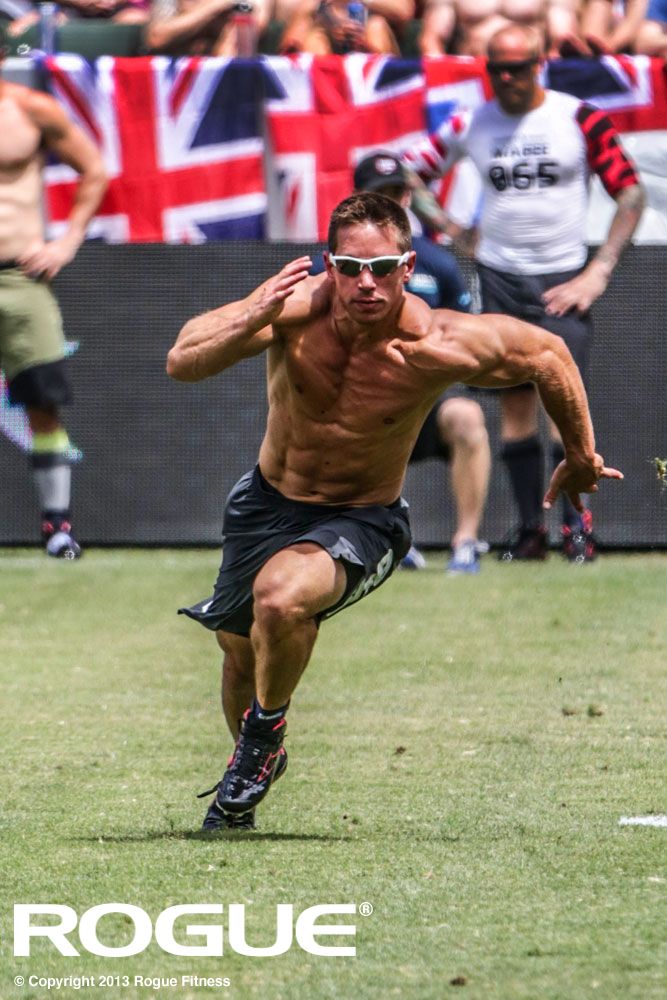 a muscular man in black shorts and a black tank top is running on a grass field. he is focused on the task at hand and pushing himself to his limits. behind him is a crowd of spectators cheering him on. the man has his shirt off, so his muscles are clearly visible. he looks determined and focused on winning. he is a true athlete! the image is taken during the 2013 rogue fitness competition.  there is a logo at the bottom of the image for rogue fitness.