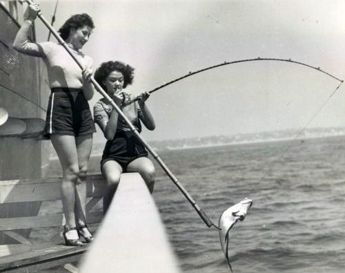 two women stand on a pier with fishing rods in hand,  one holding a  fish on the end of her line.