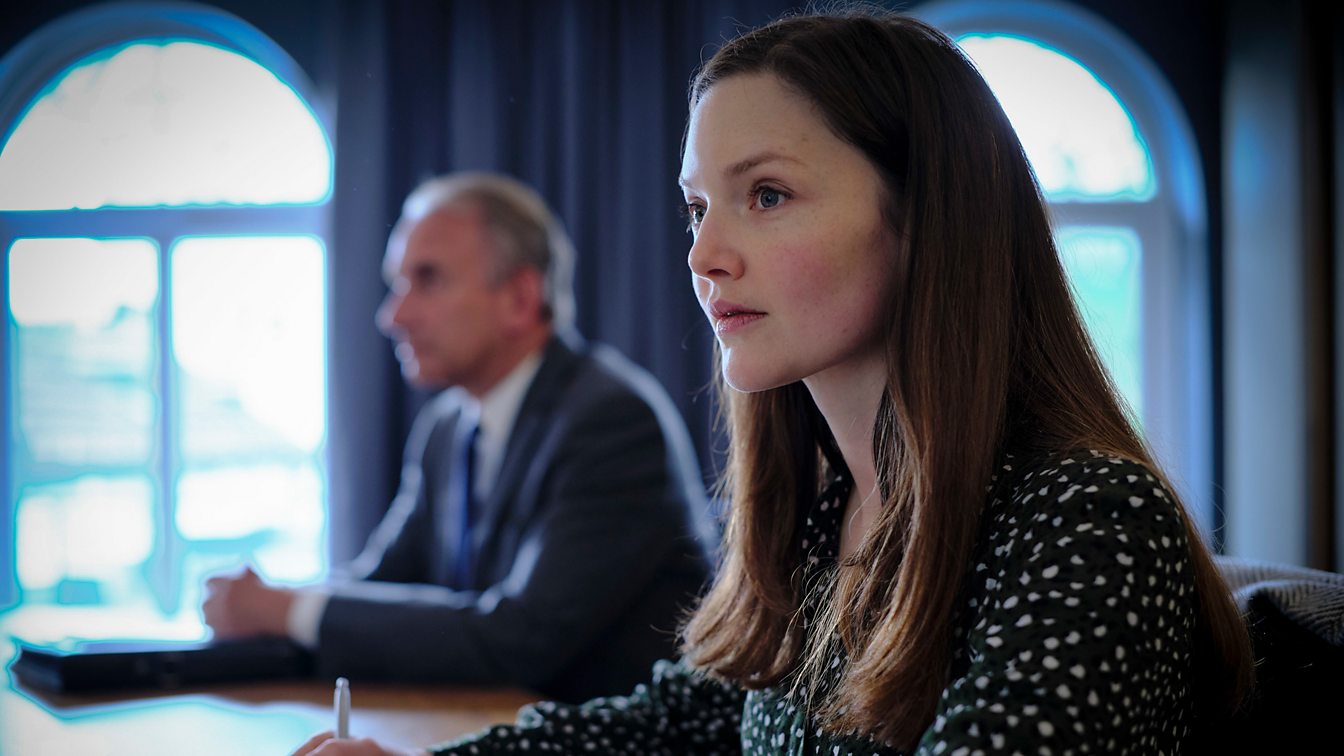 closeup of a young woman with long brown hair, sitting at a table in an office setting.  she has a serious, thoughtful expression on her face. a blurred man in a suit is visible in the background, seemingly engaged in a meeting or conversation. the setting appears professional and corporate, with large windows providing natural light.  the woman's attention is focused on something in her hands, possibly a pen and paper. the image's focus is primarily on the woman, with the background subtly out of focus.