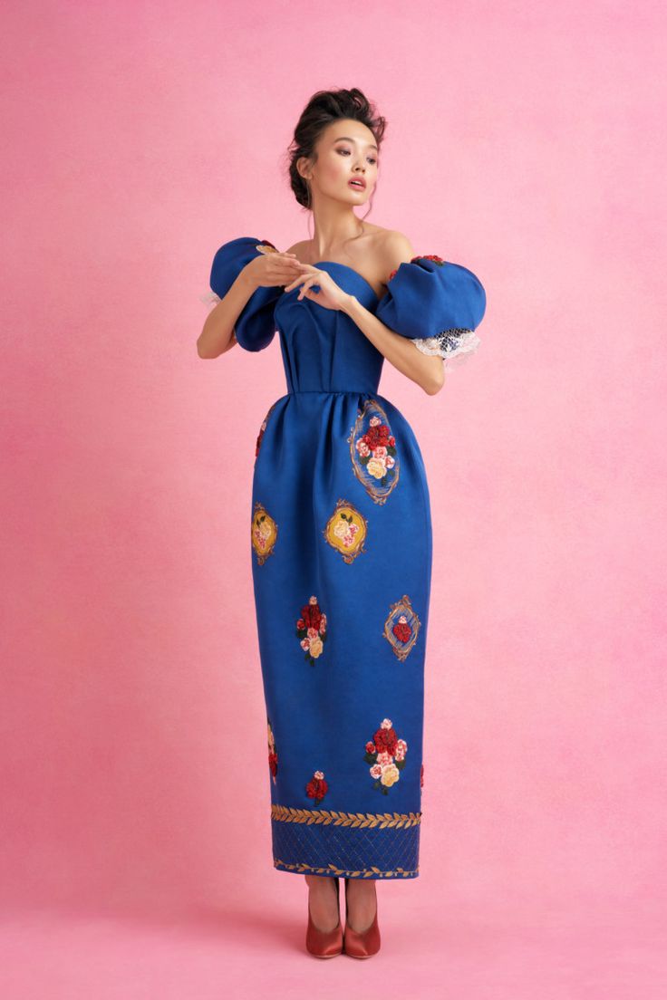 a fulllength studio shot of a fashion model wearing a beautiful blue maxi dress. the dress features puff sleeves, an offshoulder neckline, and intricate floral embroidery in red, yellow, and gold. the model has her hair styled up and is posing gracefully against a solid pink background. she is wearing red high heels. the overall image is sophisticated and elegant, highlighting the beauty of the dress and the model.