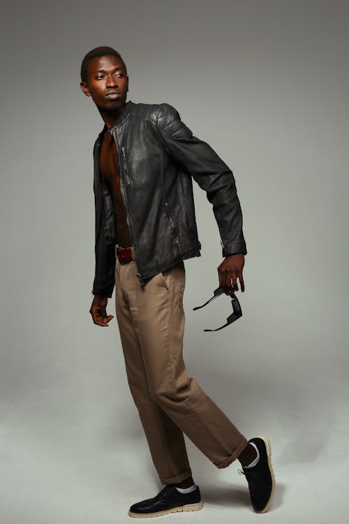 a stylish african american male model poses in a black leather jacket, brown pants, and black shoes against a gray background. he is holding a pair of sunglasses in his hand. the image is a studio shot, and the model is displaying a confident and cool demeanor. the overall aesthetic is casual yet stylish, showcasing the model's fashion sense.