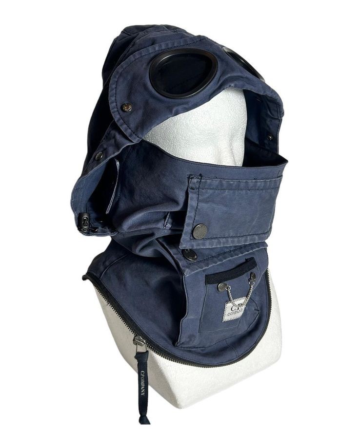 a navy blue c.p. company balaclava with goggles displayed on a white mannequin head. the balaclava features a zippered opening at the bottom and a small pocket with a logo detail.