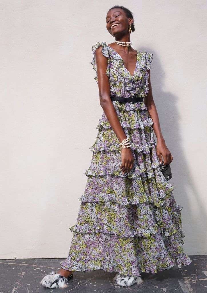 a fulllength shot of a darkskinned model wearing a beautiful tiered floral maxi dress. the dress is light purple and green with a ruffle detail on the neckline and tiers around the skirt. the model is smiling and accessorized with a black belt, pearl necklace, earrings, bracelets, and fur slides. she is holding a small black handbag.  the background is a plain lightcolored studio backdrop.