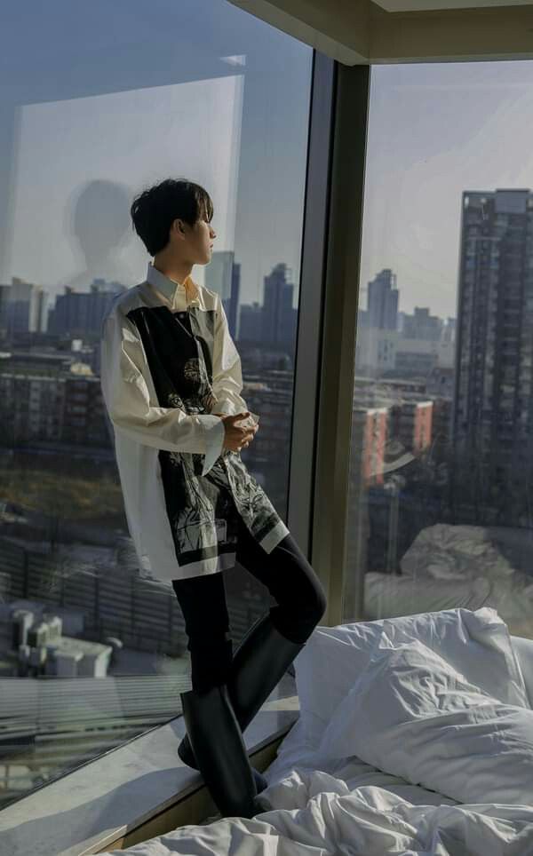 a young man stands pensively in a modern hotel room, his back to the camera as he gazes out a large window at the sprawling cityscape. he is stylishly dressed in a white overshirt layered over a black graphic tee, black pants, and tall black boots.  natural daylight streams in through the window, illuminating the room and casting a soft glow on the subject.  a portion of a white bed is visible in the foreground, hinting at the comfortable surroundings.