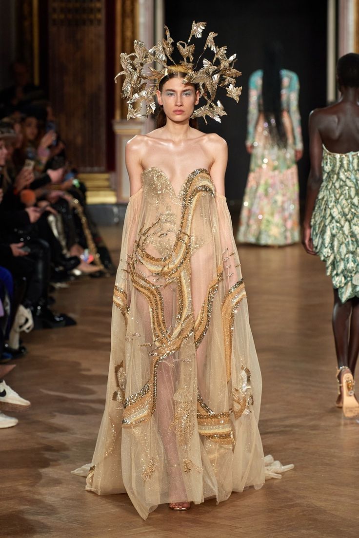 a model walks the runway in a couture gown during a fashion show. the gown is made of a sheer, goldcolored fabric and features intricate embroidery. the model is also wearing a large, gold headpiece. an audience is seated on the left side of the frame, and other models wearing similar dresses are visible in the background. the setting appears to be an elegant event space with ornate architectural details.
