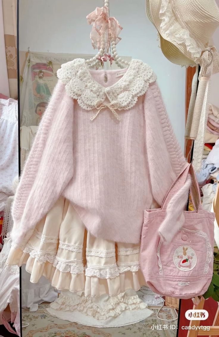 a pink and white outfit hangs against a white wall. the outfit includes a pink fuzzy sweater with a white lace collar, a long cream skirt with layers of ruffles and lace trim, and a pink tote bag with a bunny embroidery. a straw hat hangs above the outfit.