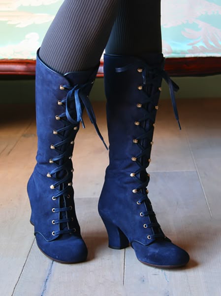 a pair of kneehigh, blue suede, laceup boots with heels are shown on a person wearing brown tights. the boots are on a lightcolored wooden floor.