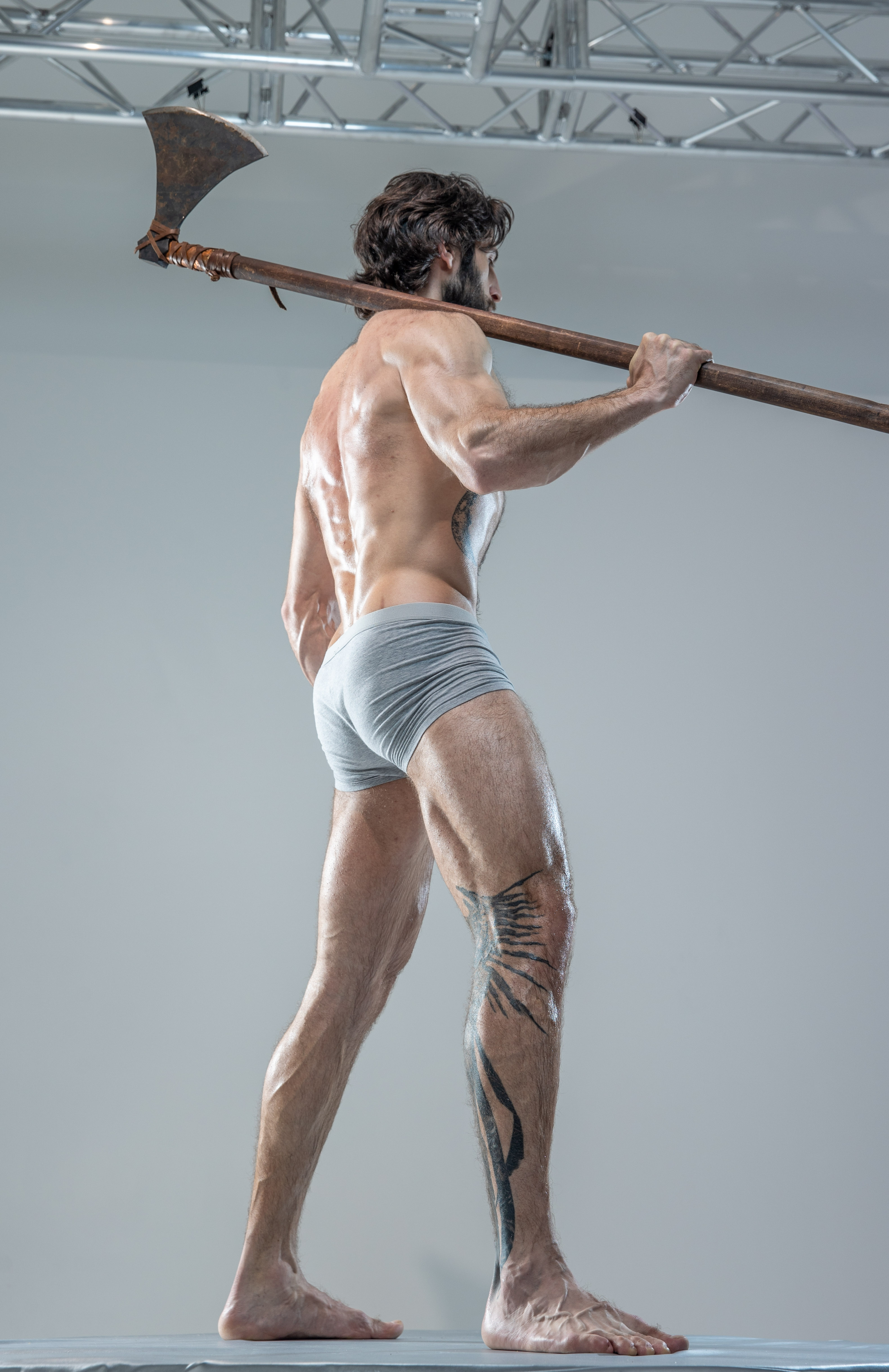 a muscular man, glistening with sweat, stands with his back to the camera, his gaze directed over his left shoulder. he holds a large, worn axe across his shoulders, his grip firm on the wooden handle. the studio setting is minimalistic, with a gray backdrop and hints of metallic scaffolding in the background.  his physique suggests strength and power, further emphasized by the primitive weapon he wields.