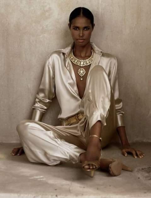 darkskinned woman, possibly a model, is elegantly posed in a seated position. she is wearing a luxurious ensemble consisting of a longsleeved satin shirt and matching trousers, both in a light beige or gold hue.  a prominent gold necklace with large stones adorns her neck, complementing the overall luxurious aesthetic. her highheeled sandals are also golden in color. the background is a textured, neutraltoned wall, providing a simple backdrop to highlight the model and her attire. the overall mood and style suggest a highfashion, editorial shoot.