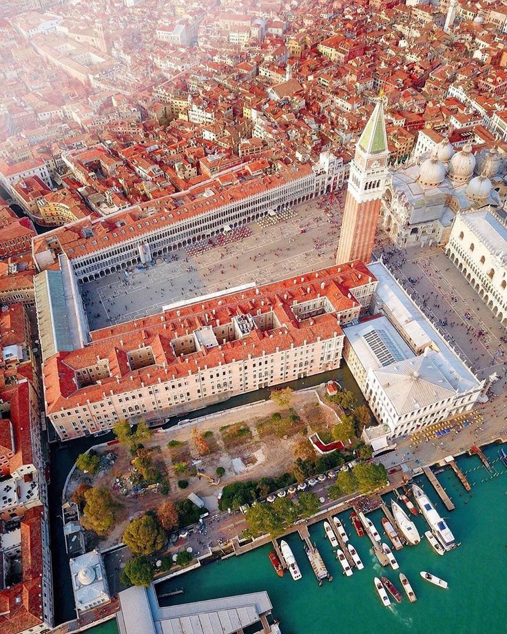 the image is a stunning aerial shot of st. mark's square in venice, italy.  the photograph showcases the iconic architecture of the square, including the campanile, doge's palace, and st. mark's basilica. the surrounding buildings, with their characteristic red terracotta roofs, extend outwards, creating a dense and visually captivating cityscape.  the canals surrounding the square are visible, with numerous boats docked along the waterfront. the overall perspective offers a unique and breathtaking view of venice's beauty and scale.  a large, empty area adjacent to the doge's palace is visible, possibly indicating ongoing construction or renovation. the green of the water and the surrounding vegetation provide a contrast to the terracotta buildings.