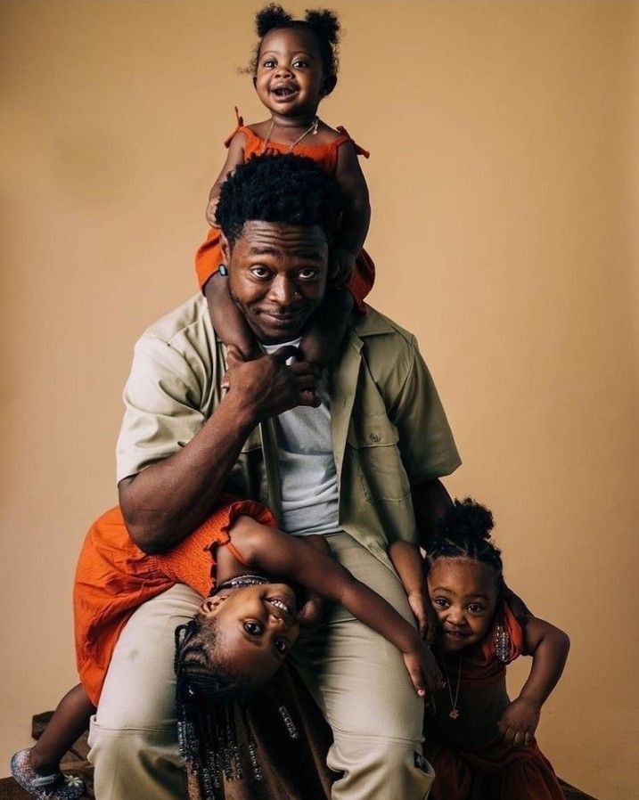 a black father is sitting in a studio with his three daughters. the father is wearing a white shirt and a khaki jacket, and the daughters are wearing red dresses. the father is holding his youngest daughter on his lap, and the other two daughters are sitting on his shoulders. all of them are smiling.