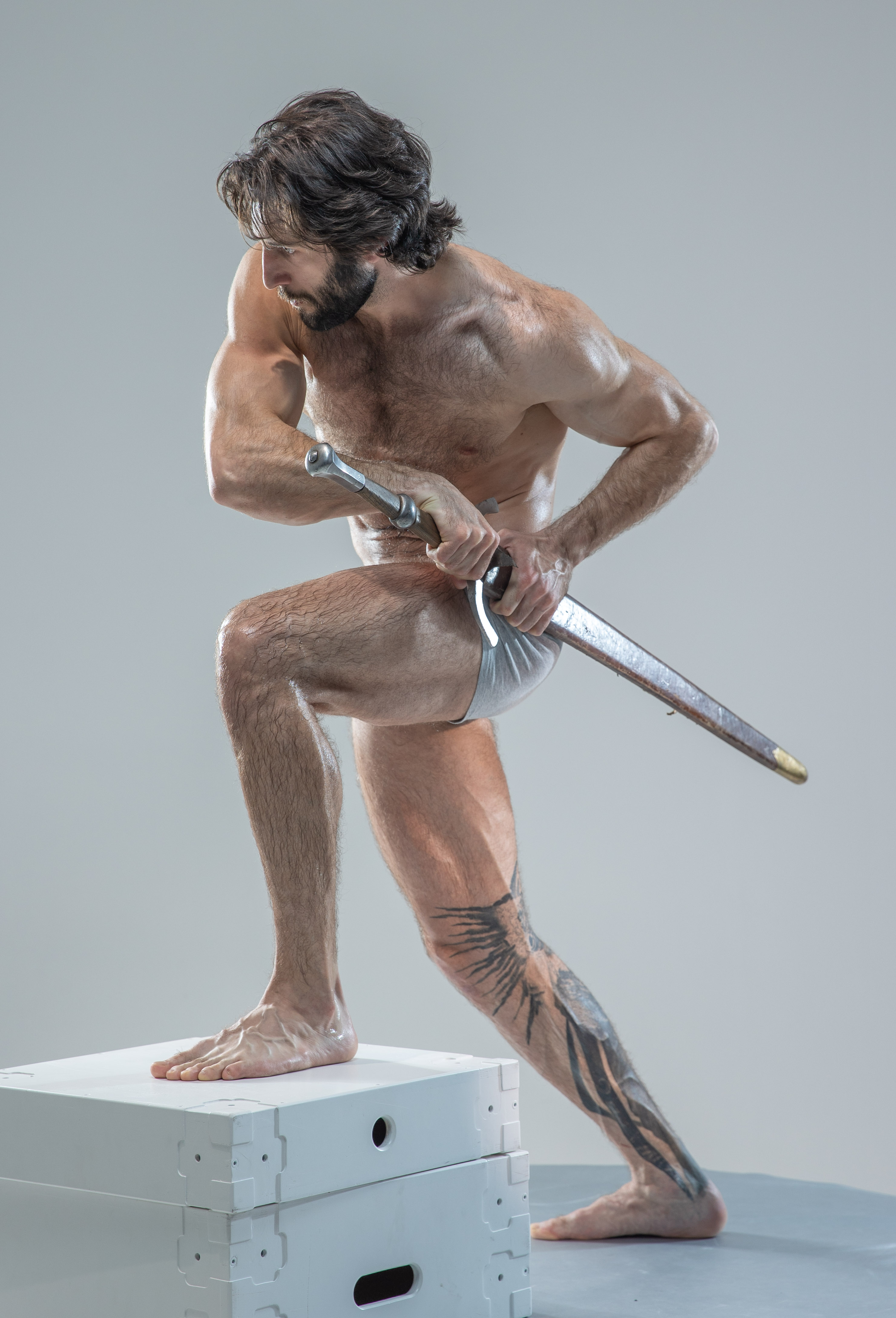 a muscular, shirtless man with long dark hair and beard stands balanced on a white box, wielding a sword. he is posed dynamically, suggesting movement and action, as if he is about to strike. he is lit with soft light against a seamless gray background, highlighting his physique. the overall aesthetic suggests classical art and warrior imagery. the man has intricate tattoos on his right leg and arm.