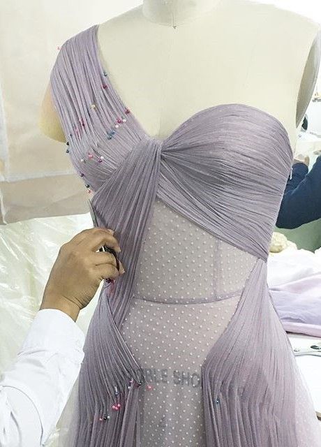 a lavender, oneshoulder dress is being designed on a mannequin. the bodice is draped with chiffon and pinned. the dress has a tulle underlay with polka dots. a hand is adjusting the drape.