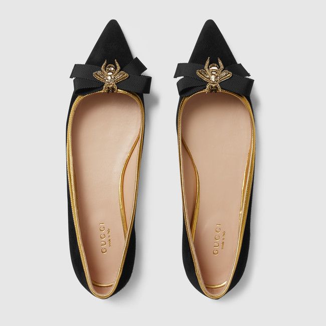 a pair of black suede gucci women's flats are shown from above.  the shoes have a pointed toe, gold trim around the edges, and a gold embroidered bee with a black bow on the top.  the gucci logo is visible inside the shoe.  the shoes are positioned against a plain grey background.