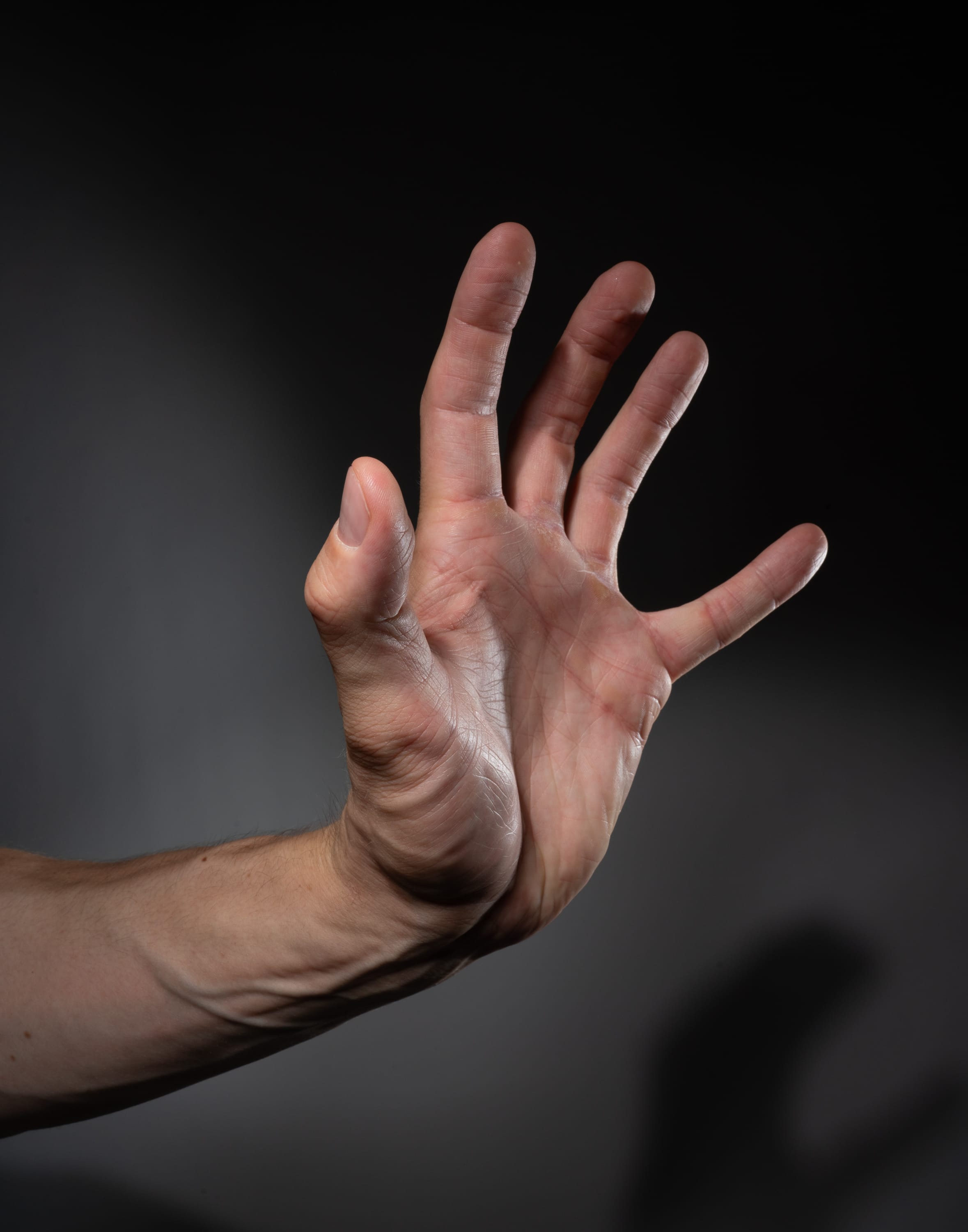 a male hand is extended in front of a grey background with dramatic lighting. the hand is in focus and positioned in the center of the image. the fingers are spread apart and the palm is facing the viewer. the hand appears to be making a stop gesture.