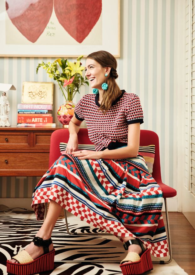 a woman with shoulderlength brown hair is sitting on a red chair. she is wearing a red and white patterned shirt and a colorful geometric patterned skirt. she is also wearing platform sandals. she is smiling and laughing. the background includes a red heart painting, striped wallpaper, and a zebra rug. there are also books and a vase of flowers on a wooden cabinet in the background.
