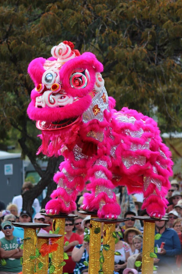 a vibrant pink and silver chinese lion dance costume is the focal point, perched atop golden poles adorned with greenery. the lion's mouth is open in a playful roar, its eyes wide and sparkly. a crowd of people of all ages watches the performance with smiles and interest. the setting appears to be an outdoor park with lush green trees in the background, bathed in bright sunlight. the overall atmosphere is joyful and celebratory.