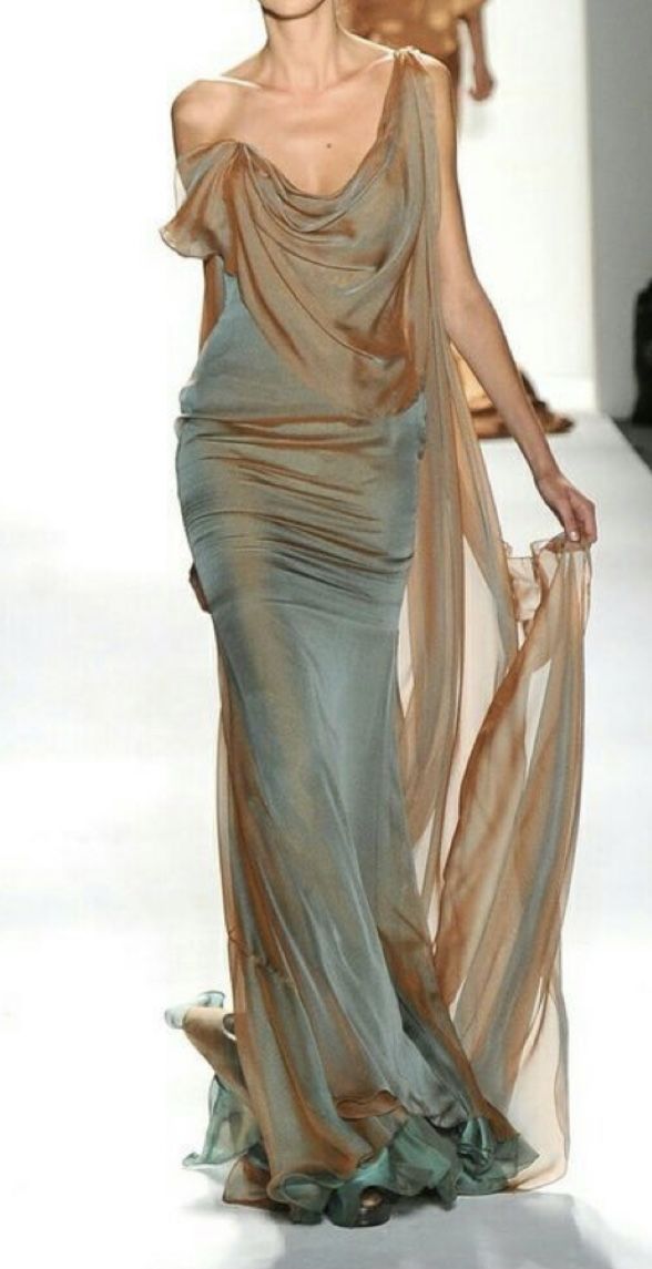 a runway model is showcasing a long, elegant gown. the dress is primarily a pale teal color, with draped bronze fabric creating a flowing, sheer overlay. the dress has a cowl neck and a fitted silhouette that flows into a long, sweeping train. the model's pose and the overall aesthetic suggest high fashion and sophistication. the background is a classic runway setting.
