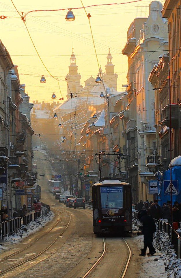 a captivating image showcasing a tram traversing a snowy street in lviv, ukraine, during a picturesque sunset. the golden hour light casts a warm glow on the historical buildings lining the street, which are characterized by their vintage architecture. the scene is serene, with a blend of urban elements and natural winter ambiance.  the tram stands out as a focal point, moving through the street with pedestrians and buildings forming a captivating backdrop. the overall atmosphere is nostalgic and peaceful, highlighting the charm of the city.