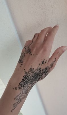 the image shows a hand and part of an arm with a detailed henna tattoo. the tattoo features intricate patterns, flowing lines, dots, and star shapes, covering the hand and extending up the arm. the hand is fairskinned and posed against a light, solid background.