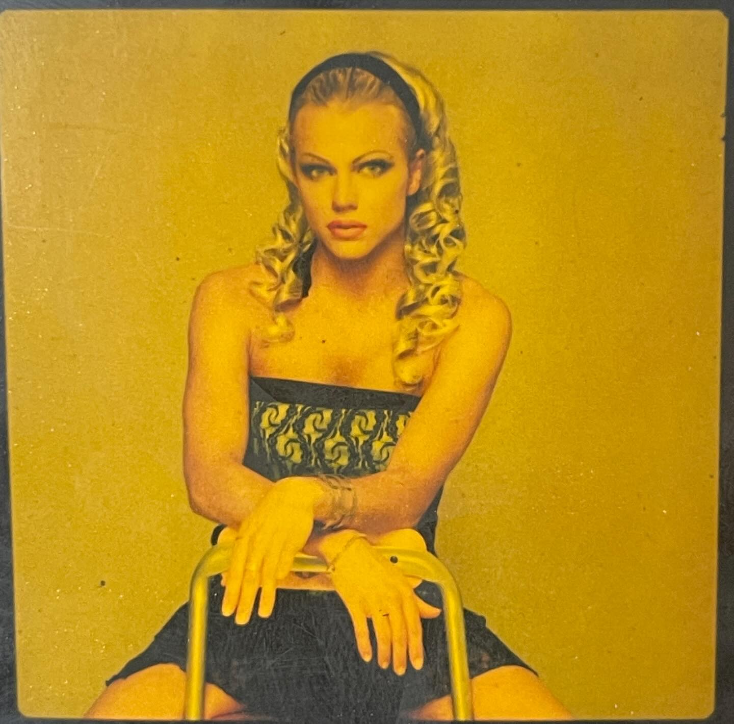 a vintagestyle portrait of a blonde woman with curly hair, wearing a black strapless dress and headband. she is sitting on a chair against a gold background and looking directly at the camera. the image has a slightly grainy, textured quality. the overall mood is glamorous and confident.