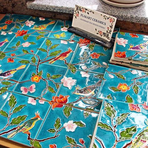 the image shows a collection of handmade ceramic tiles arranged on a surface. the tiles are square and feature a vibrant turquoise or teal background with a repeating pattern of stylized birds and flowers in bright, rich colors. the design appears to be inspired by persian or turkish motifs. a sign reading "plakart ceramics, handmade tile design studio" is visible in the upper middle of the image, indicating the origin of the tiles.  several stacks of the tiles are visible, indicating they are for sale or display. the overall impression is one of artistry, craftsmanship, and vibrant color.