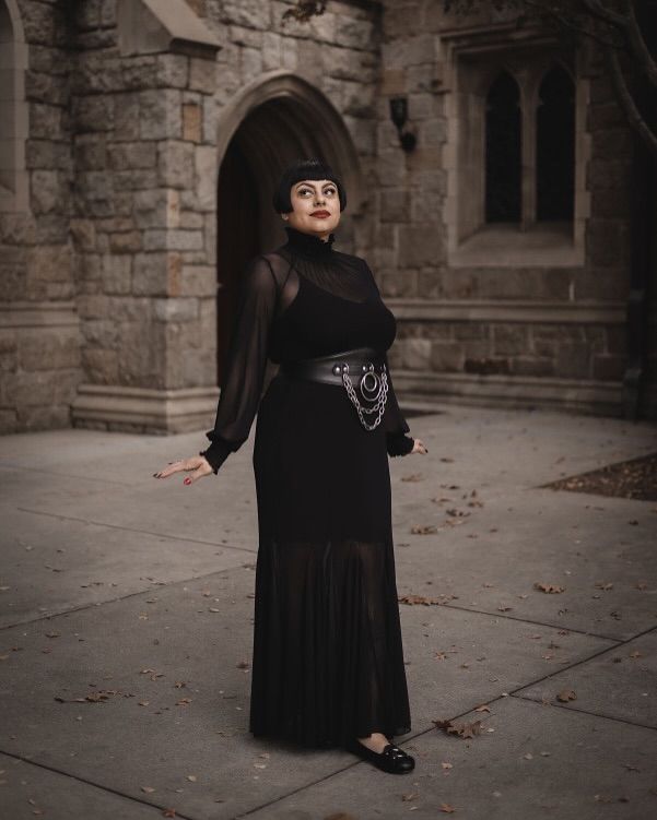 a woman in a long, black gothicstyle dress stands outdoors in front of a stone building. the dress is sheer at the bottom and features a pleated skirt and a black chain belt.  she is looking upward, and her expression is serene.  fallen leaves are scattered on the ground around her. the overall atmosphere is dark and elegant.